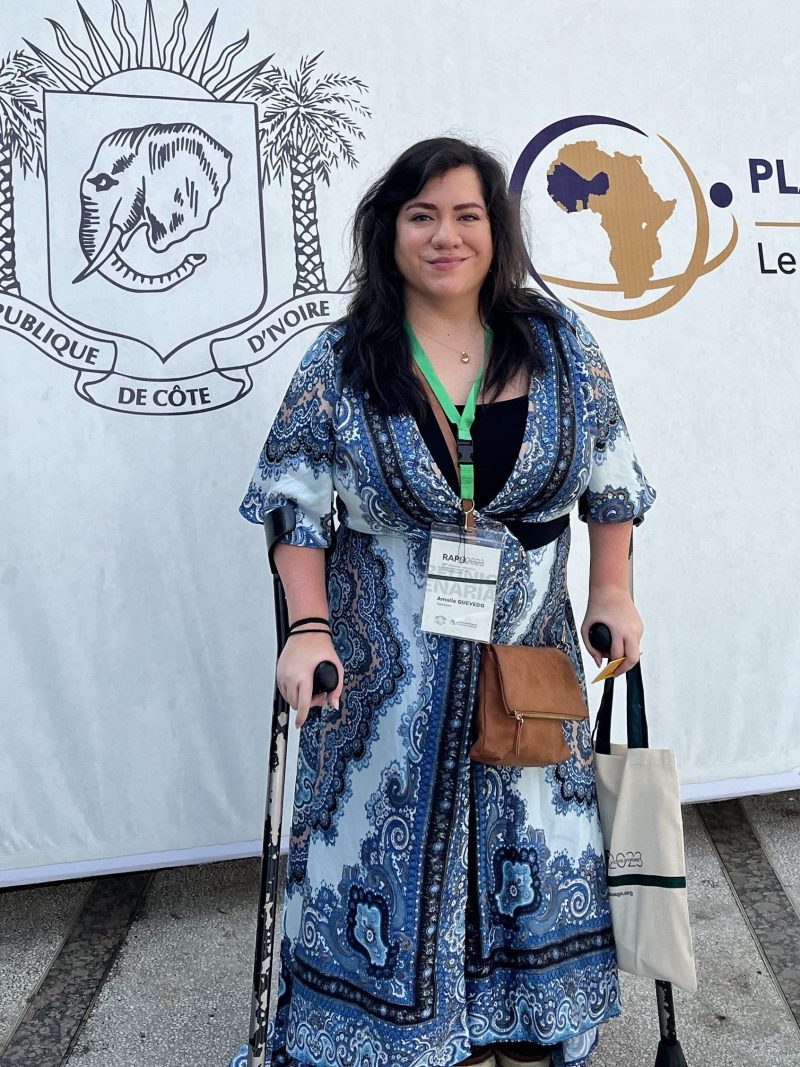 A photo of Amalie Quevedo standing in front of a banner with the coat of arms of Ivory Coast which includes the profile of an elephant, a sunrise and two palms. The banner also has a map of Africa, highlighting west Africa. Amalie is smiling towards the camera with long brown hair, wearing a blue and white wrap dress and a conference tag on a green strap around her neck. She is supporting herself on two crutches.