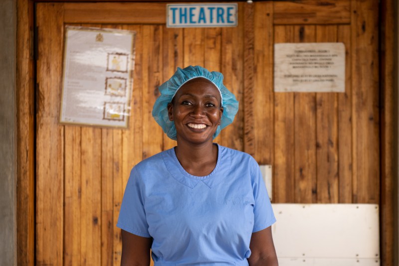 Photo of Dr Moira Chinthambi wearing a typical surgeon's outfit, blue scrubs and a hair cap. She is smiling and looking into the camera. Behind her is a sign with the word THEATRE.