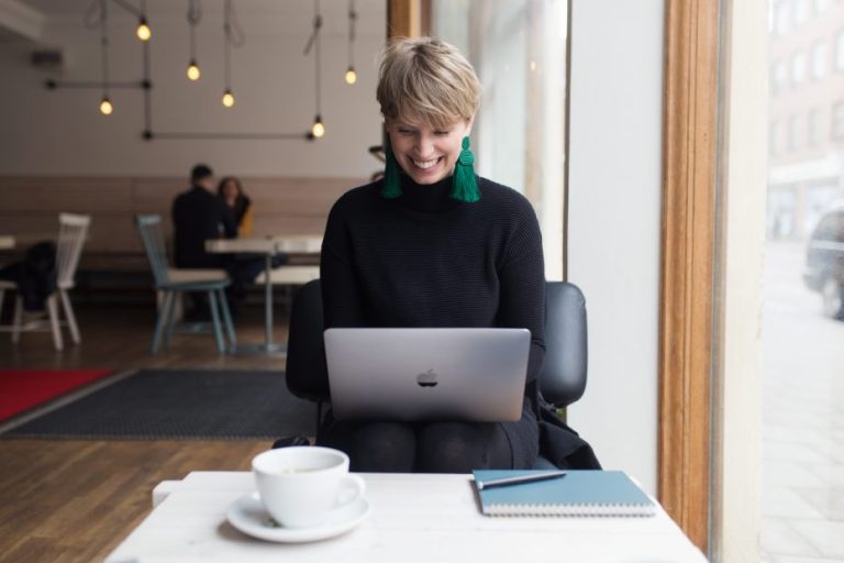 Photo of Julia working with her computer at a café. She has short blonde hair, wearing big green earrings, writing on a laptop. On the table in front of her is a cup of tea and a notebook and pen.