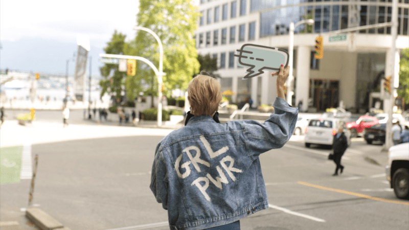 Photo of Julia Wiklander, her back with a jeans jacket with the letters GRL PWR on the back, and holding a sign in the image of a speech bubble.