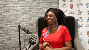 Dr. Monica Mhoja speaks with Girls' Globe about women's land rights and the Stand 4 Her Land campaign in the Girls' Globe Solidarity Studio. Dr. Monica Mhoja is speaking into a podcast microphone, wearing a red short sleeved dress, a blue and multi-colored beaded necklace and matching earrings.