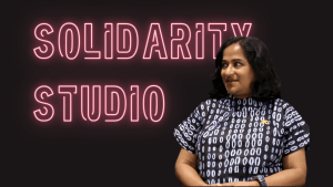 Neon pink Text with the words Solidarity Studio on a black backround and Deepti Mathew in a blue and white batik blouse.