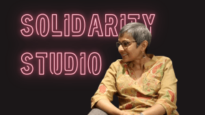 Pink neon text on Black background with the words SOLIDARITY STUDIO and the profile of Lopa Banerjee in a yellow tunic.