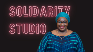 The profile of Mwanahamisi Singano in front of a black background with pink neon words Solidarity Studio. Mishy wears a turquoise headscarf, black glasses and a blue tie die dress. She smiles at the camera.