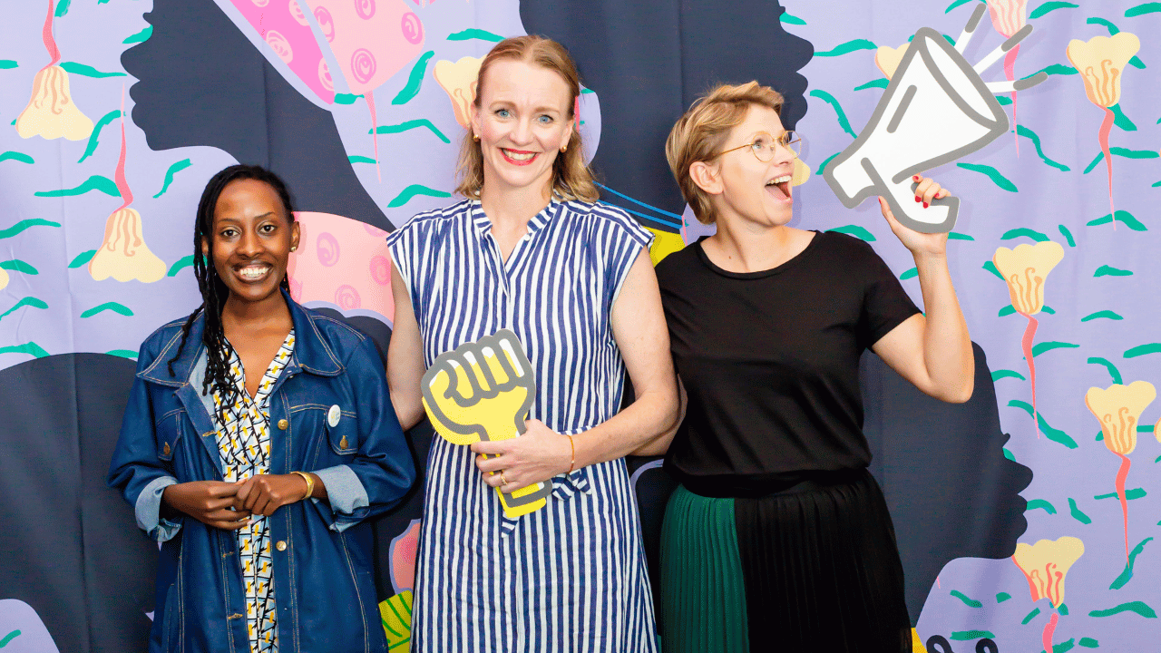 Amina Umuhoza, Margaret Butler and Julia Wiklander outside of the Solidarity Studio. They are standing in front of a large illustration of the portraits of women.
